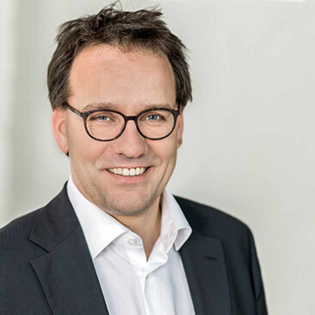 Mann mit Brille und Anzug l&auml;chelt freundlich in neutralem Hintergrund.
