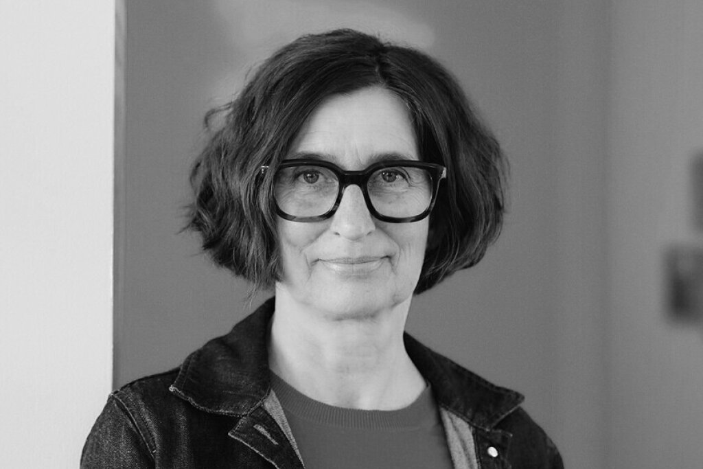 Frau mit kurzer, lockiger brauner Haarfrisur tr&auml;gt eine blaue Bluse und eine Jeansjacke vor roter Wand.