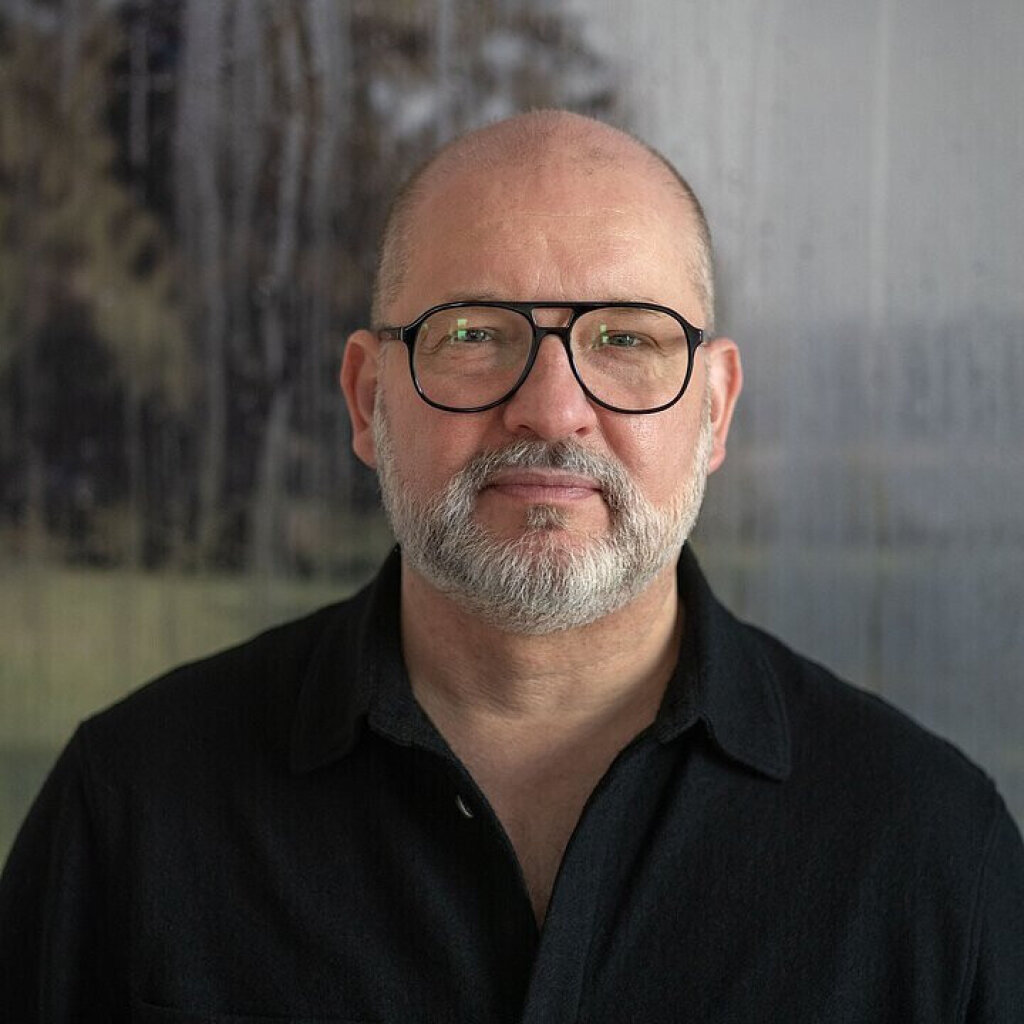 Mann mit Brille und Bart, tr&auml;gt ein schwarzes Hemd, Hintergrund zeigt unscharfe B&auml;ume.
