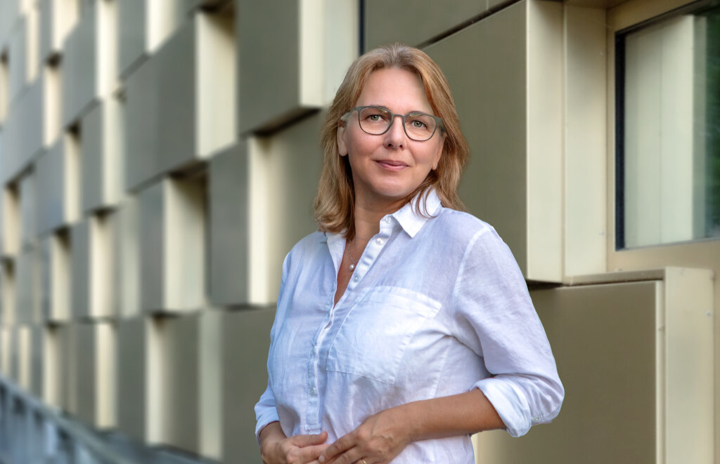 Frau mit Brille und halblangem Haar steht in halbnaher Einstellung vor einer Reliefwand.