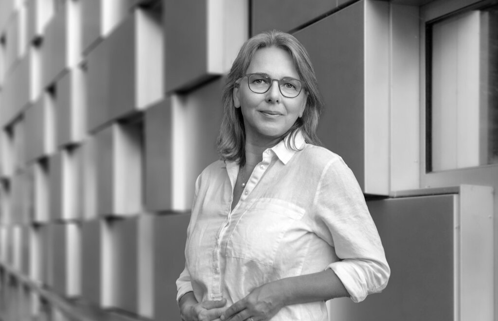 Frau mit Brille und halblangem Haar steht in halbnaher Einstellung vor einer Reliefwand.